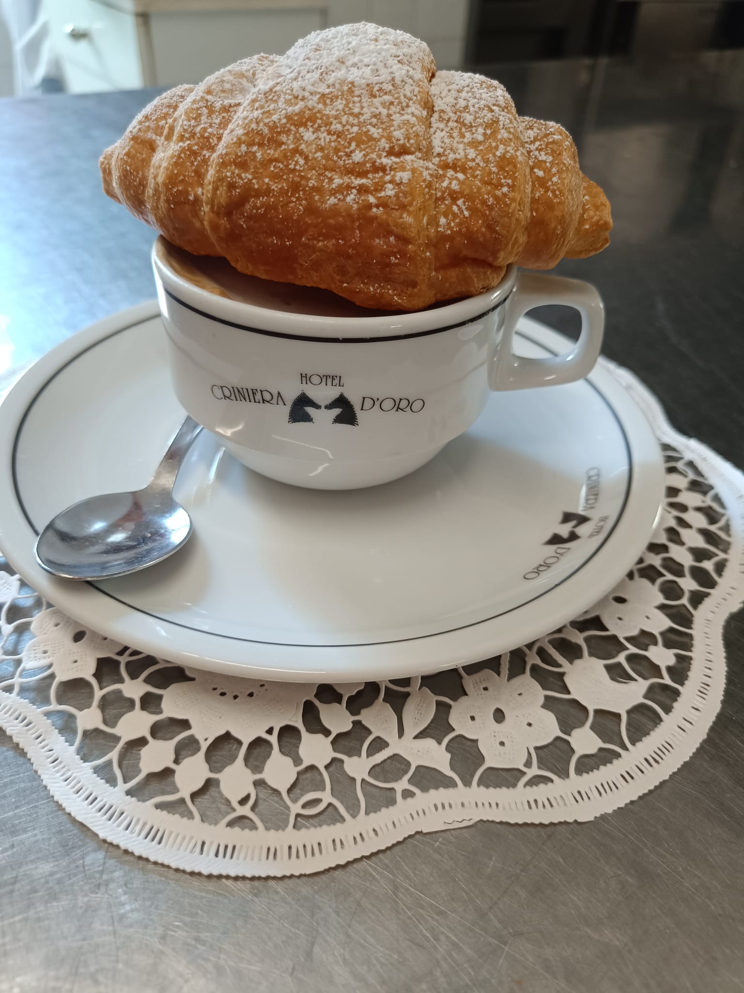 Cornetto spolverato di zucchero a velo su una tazza di cappuccino con il logo Hotel Criniera d'Oro